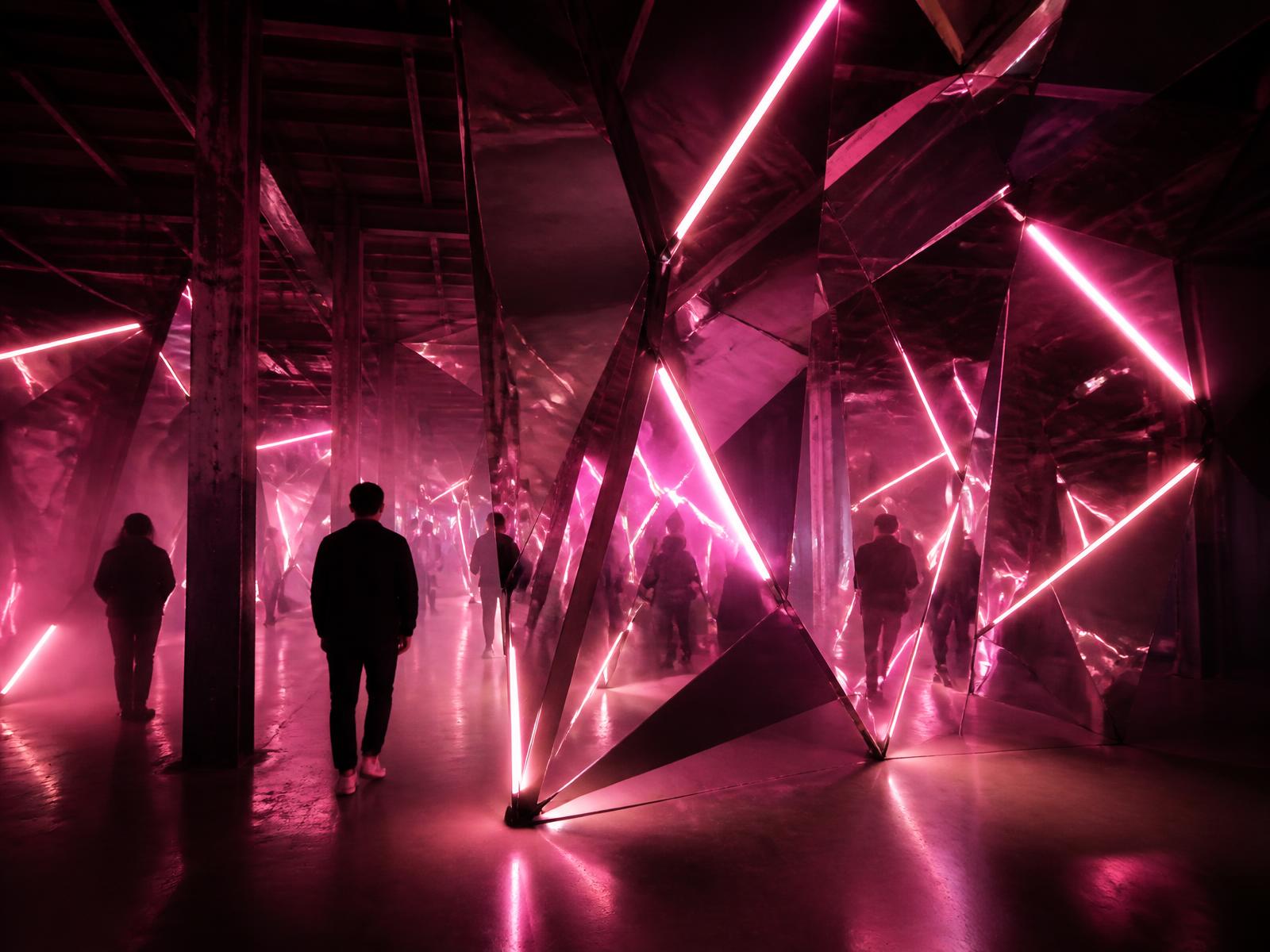 Instalación inmersiva con estructuras espejadas y neón rosa
